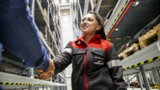 A smiling Linde MH service technician shakes hands with another person between warehouse shelves.