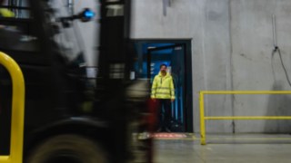 A man in safety clothing steps out of a door into a factory hall. A forklift truck drives past. A man in safety clothing steps out of a door into a factory hall. A forklift truck drives past.