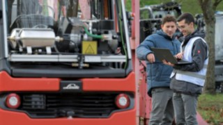 Two men stand in front of an autonomous forklift and look at a laptop together