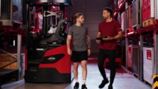Two people in sportswear standing in front of a Linde reach truck Ri16, one holding a tablet.