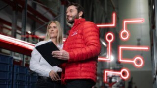 A man and a woman are standing in a warehouse, each holding a clipboard. 