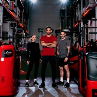 A woman and two men in athletic wear are standing in a warehouse surrounded by Linde Material Handling vehicles.