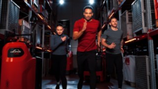 A woman and two men in athletic clothing are walking down a warehouse aisle.