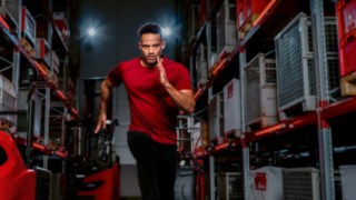A man in athletic clothing runs toward the camera between warehouse shelves.