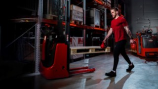 A man in athletic clothing is carrying dumbbells down a warehouse aisle.