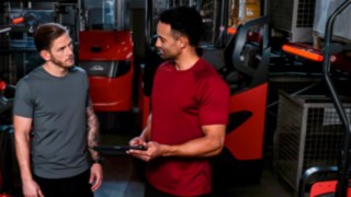 Two men in athletic wear are standing in a warehouse, talking.